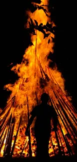 A silhouette stands before a towering bonfire at night.