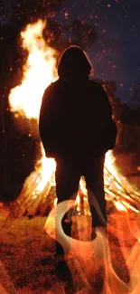 Silhouetted figure standing before a large bonfire at night.