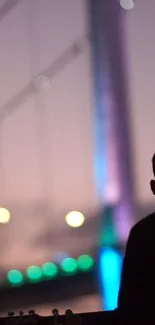 Silhouette of a man with guitar by colorful lit bridge at sunset.