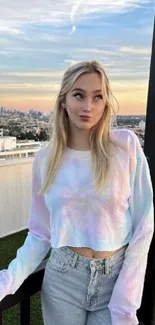 Model on a rooftop with city skyline at sunset, wearing a pastel top and denim.