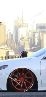 White car with red wheels in front of a city skyline at sunset.