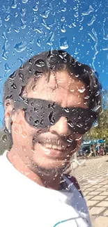 Smiling man behind raindrop-covered glass outdoors.