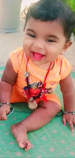 Smiling baby in orange outfit on green mat outdoors.