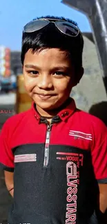 A young boy smiles in a red shirt with sunglasses, set against a bright urban backdrop.