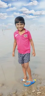 Smiling child standing by water under a blue sky with clouds.