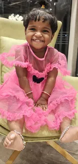 Smiling child in a pink dress sitting on a green chair indoors.