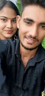 Smiling couple taking a selfie outdoors, with greenery in the background.