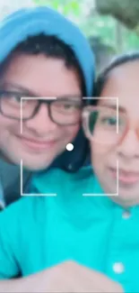 Two friends taking a smiling selfie in a natural setting, both wearing glasses.