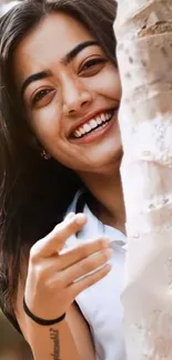 A cheerful young woman smiling behind a tree, exuding joy and positivity.