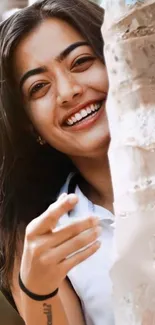 A smiling woman peeks from behind a tree in warm natural light.