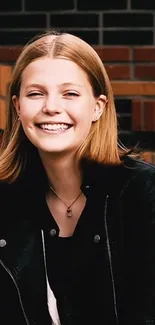 Smiling woman in a leather jacket against a brick wall.