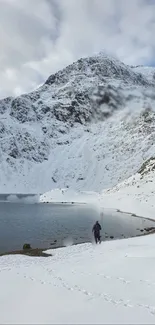 A lone figure walks through a snowy mountain landscape beside a calm lake.