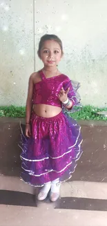 Girl in a pink sparkly dress stands smiling against a wall.