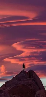 Lone figure on rocky peak under vibrant orange sunset clouds.