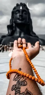 Hand with beads and statue under cloudy sky.