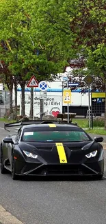 Black sports car with yellow stripe on a tree-lined road.