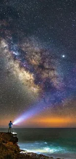 A person stargazes at the Milky Way on a rocky coastline under a dark blue sky.