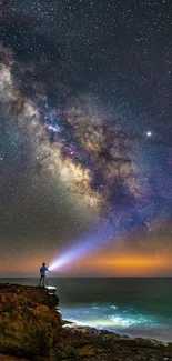 Man with flashlight under star-filled sky by ocean.