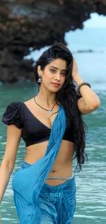 Woman in a blue sari standing by the ocean on a rocky beach.