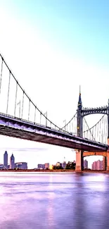 Suspension bridge over river at sunset with city skyline.