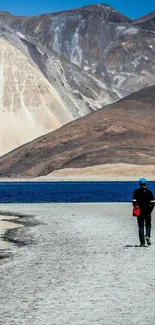 Person walking by blue lake against mountain backdrop.