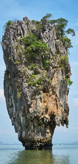 Tall rock island with lush trees in clear waters under a blue sky.