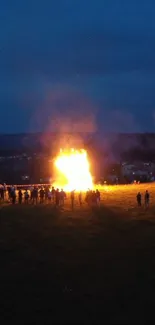 A large bonfire under a deep blue night sky with silhouettes of gathered people.