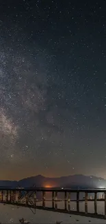 Starry night sky over mountains with a bridge in view.