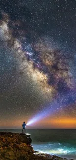 Person holding light under starry Milky Way sky over ocean.