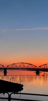 Sunset over silhouetted bridge with serene water reflections.