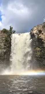 High waterfall with rainbow in rocky landscape under blue sky.