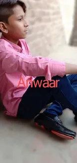 Boy in pink shirt sitting against a brick wall, looking thoughtful.