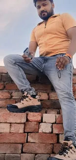 Man in jeans and orange shirt posing on a brick wall.