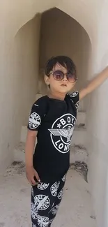 Child in black outfit with sunglasses in a white corridor.