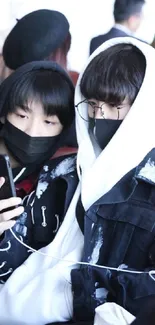 Two stylish individuals in hoodies and masks, viewing a phone.