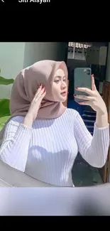 Chic woman in hijab takes a stylish mirror selfie with serene background.