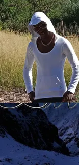 Person in white hoodie amidst nature with mountain backdrop.