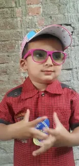 Child wearing pink glasses and red shirt against a brick wall.