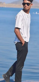 Man in stylish outfit walking by a lake with mountains in background.