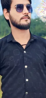 Man in a patterned black shirt with sunglasses against a scenic backdrop.