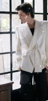 Man in a white blazer stands by a window in a stylish indoor setting.