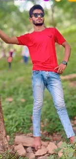 Man in red shirt and jeans leaning against a tree in a green forest setting.