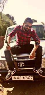 Man in red plaid shirt sitting on car hood, holding a stick.