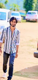 Man in striped shirt outdoors with cars parked behind on a sunny day.