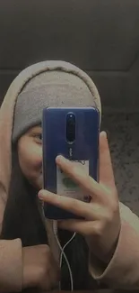 Person wearing hoodie and beanie taking a selfie in a mirror with a phone.