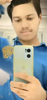 Young man taking selfie in mirror with blue shirt and smartphone.