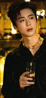 Man in black shirt and gold necklace with a drink, bar background.