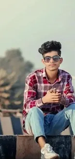 Young man in plaid shirt and sunglasses sitting outdoors.