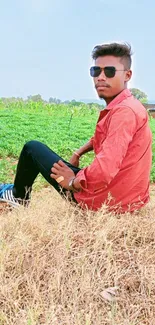 Young man in red shirt sitting outdoors with a green field backdrop.