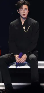 Stylish performer in black suit on stage, seated elegantly.